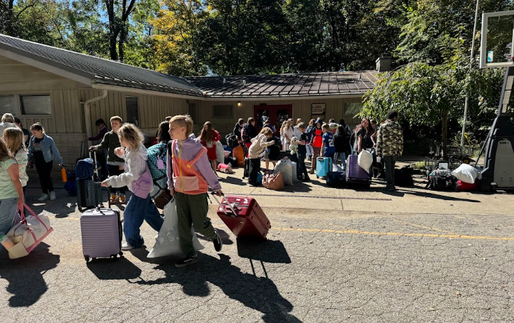 A group of children and adults walking with rolling suitcases and bags in front of a camp building.