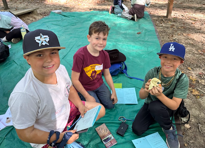 Three young children smile at the camera while sitting on a large green tarp outdoors, holding booklets and a small animal skull.