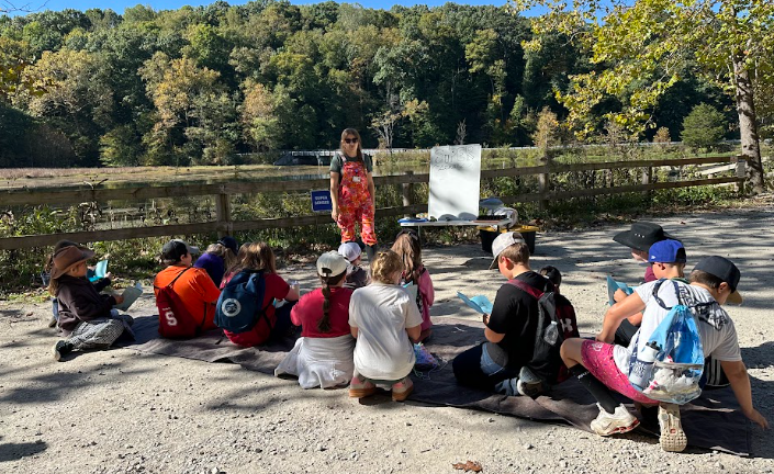A group of children sit on a tarp outdoors, facing an instructor standing near a whiteboard.
