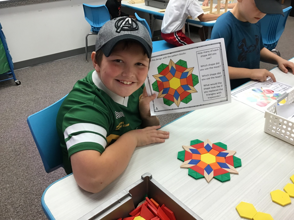 A smiling child holds up a colorful pattern block design paper in a classroom, with the matching wooden block creation on the table.