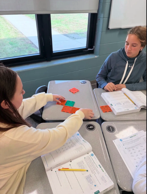 Two students sit at individual desks in a classroom, working on an assignment with colorful magnetic tiles.