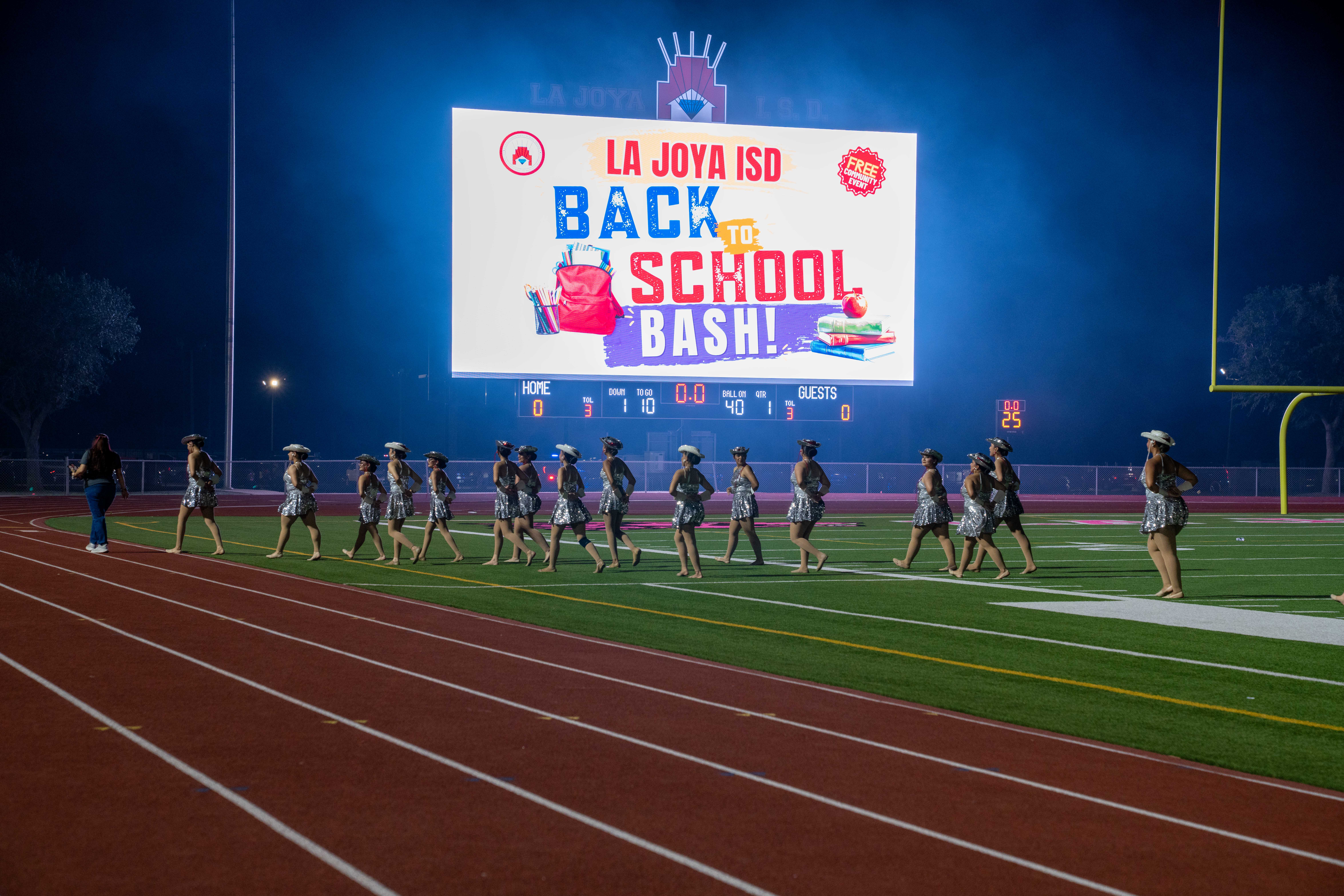 A group of dancers in sparkly outfits walk across a football field at night, with a large screen displaying 'LA JOYA ISD BACK TO SCHOOL BASH!'