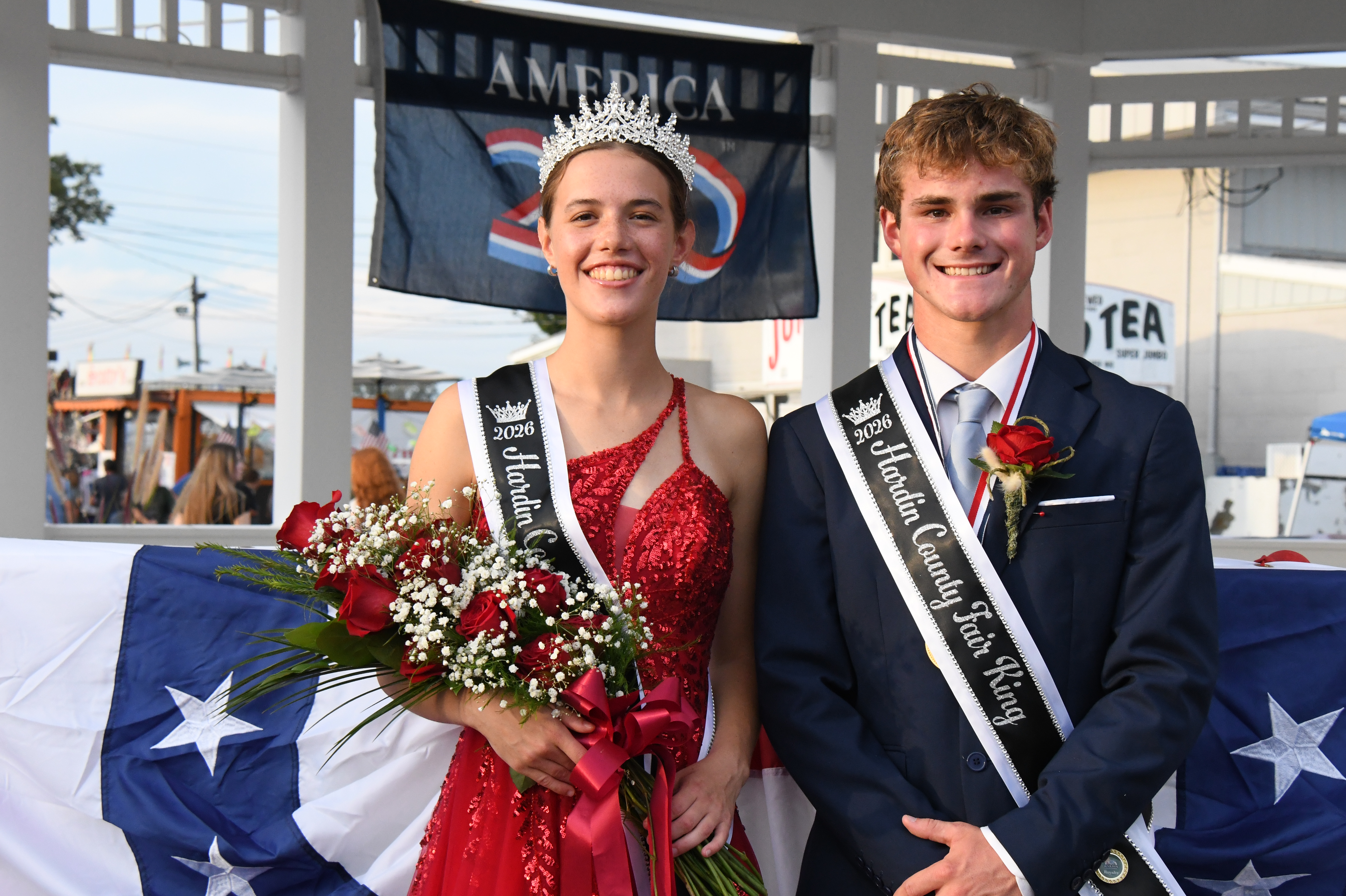 A young woman in a sparkling red dress and a crown holds a large bouquet of roses, standing next to a young man in a suit with a sash that reads "Hardin County Fair King".