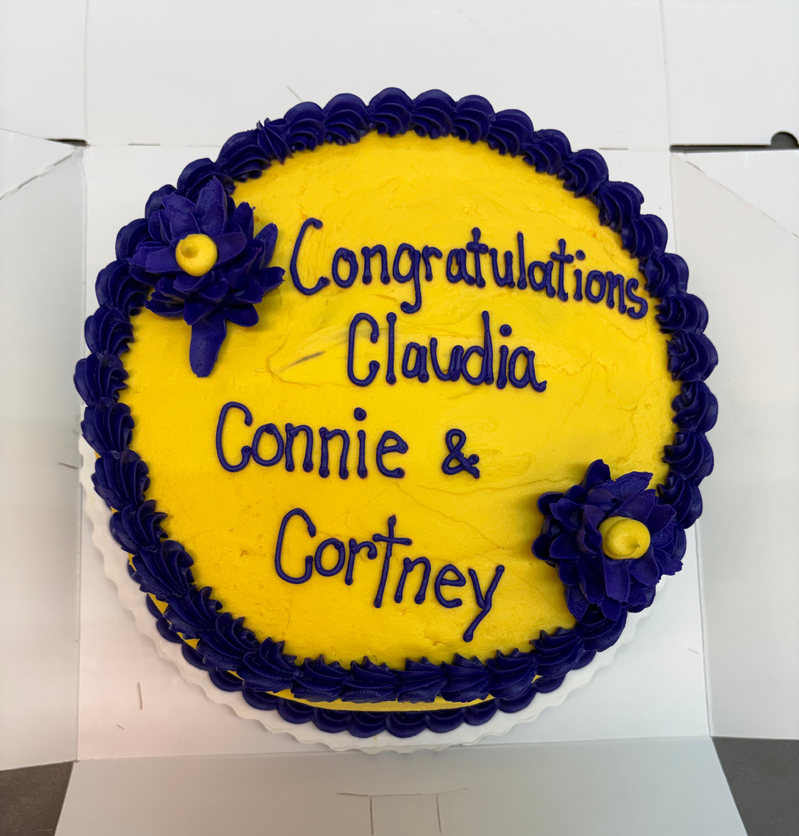 A yellow cake with purple frosting reads 'Congratulations Claudia, Connie & Cortney'.