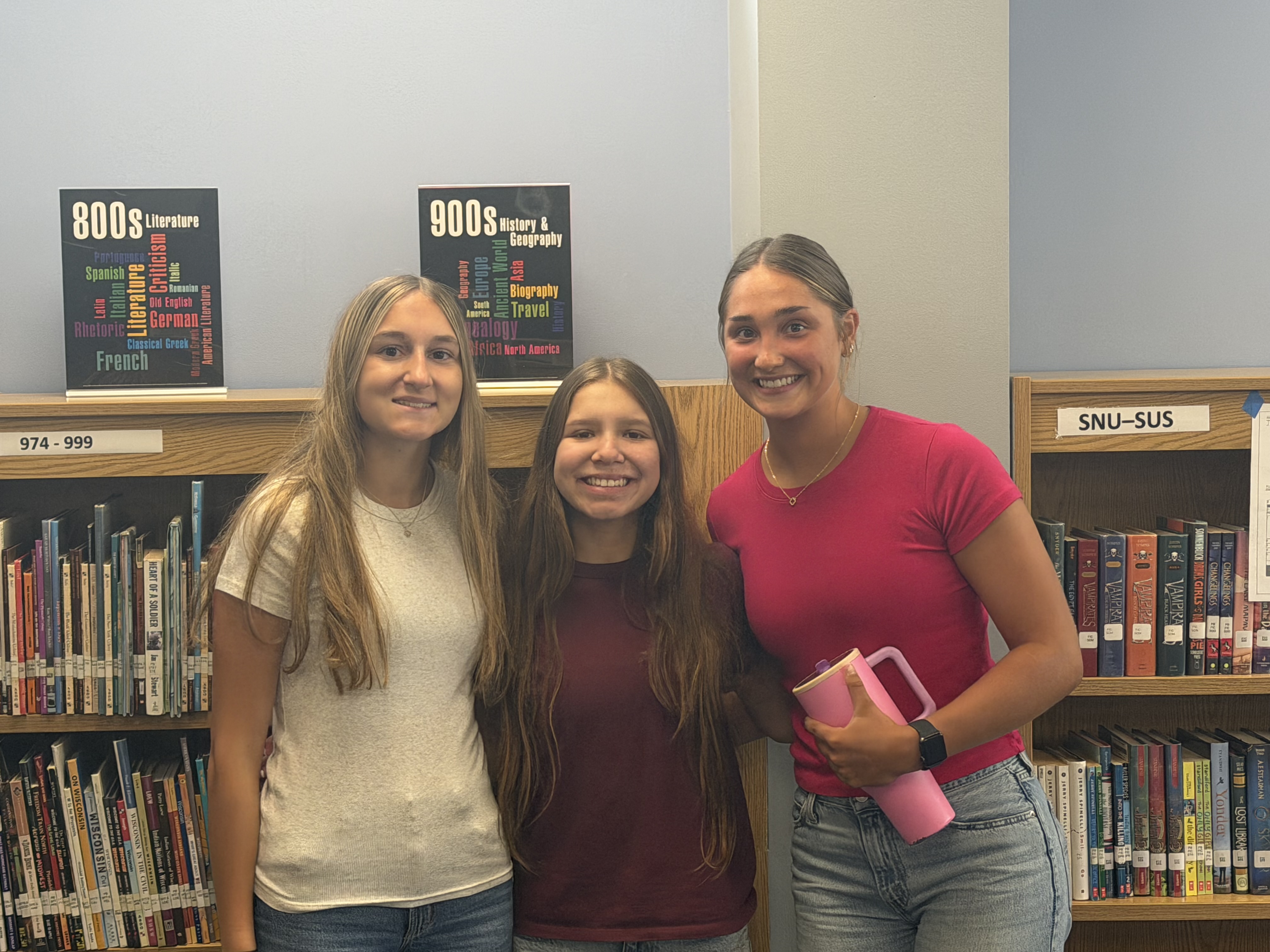 Three smiling young women stand in front of bookshelves in a library, with educational posters visible behind them.
