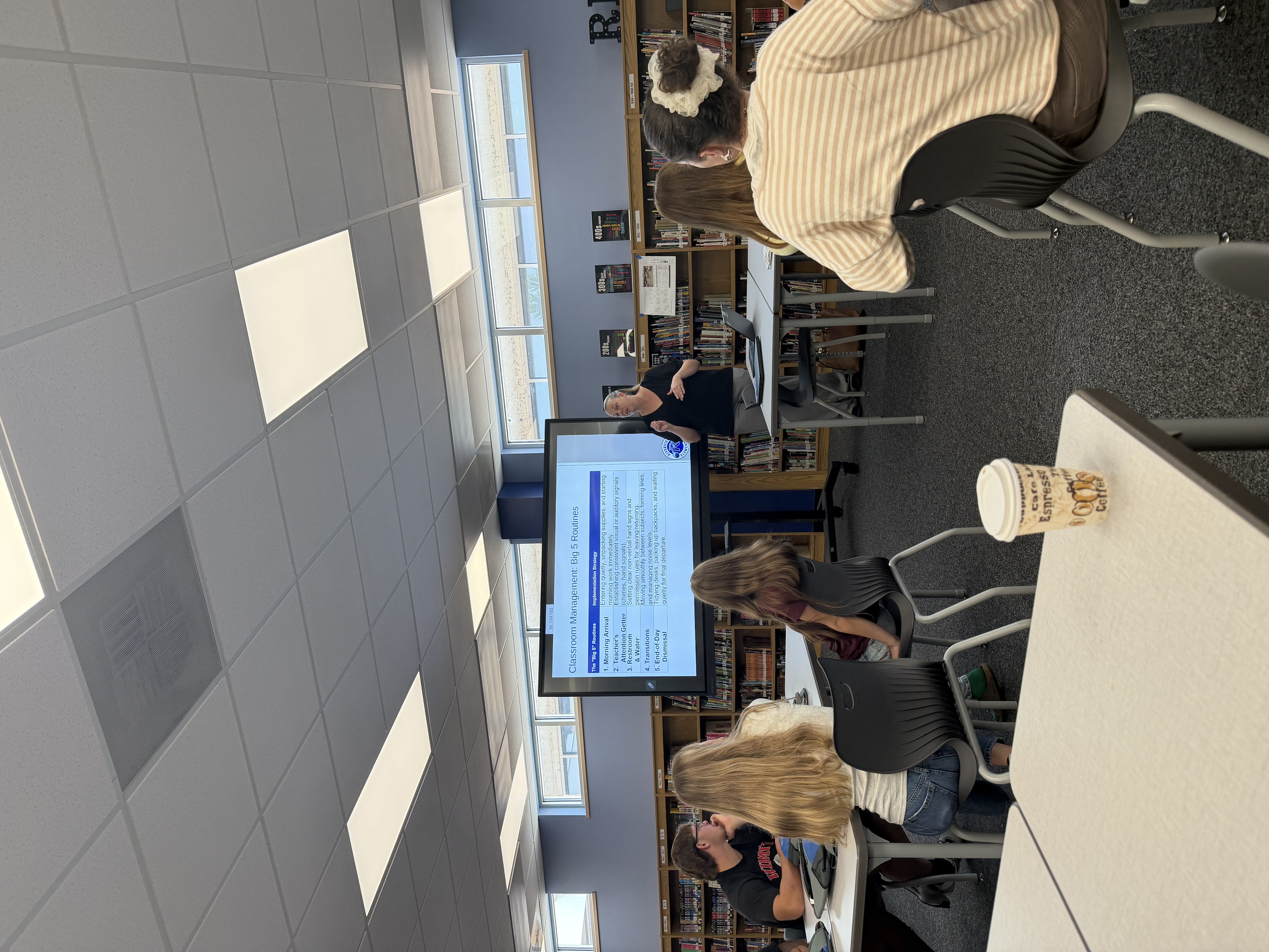 A teacher presents a slide on "Classroom Management: Big 5 Routines" to students in a library setting.