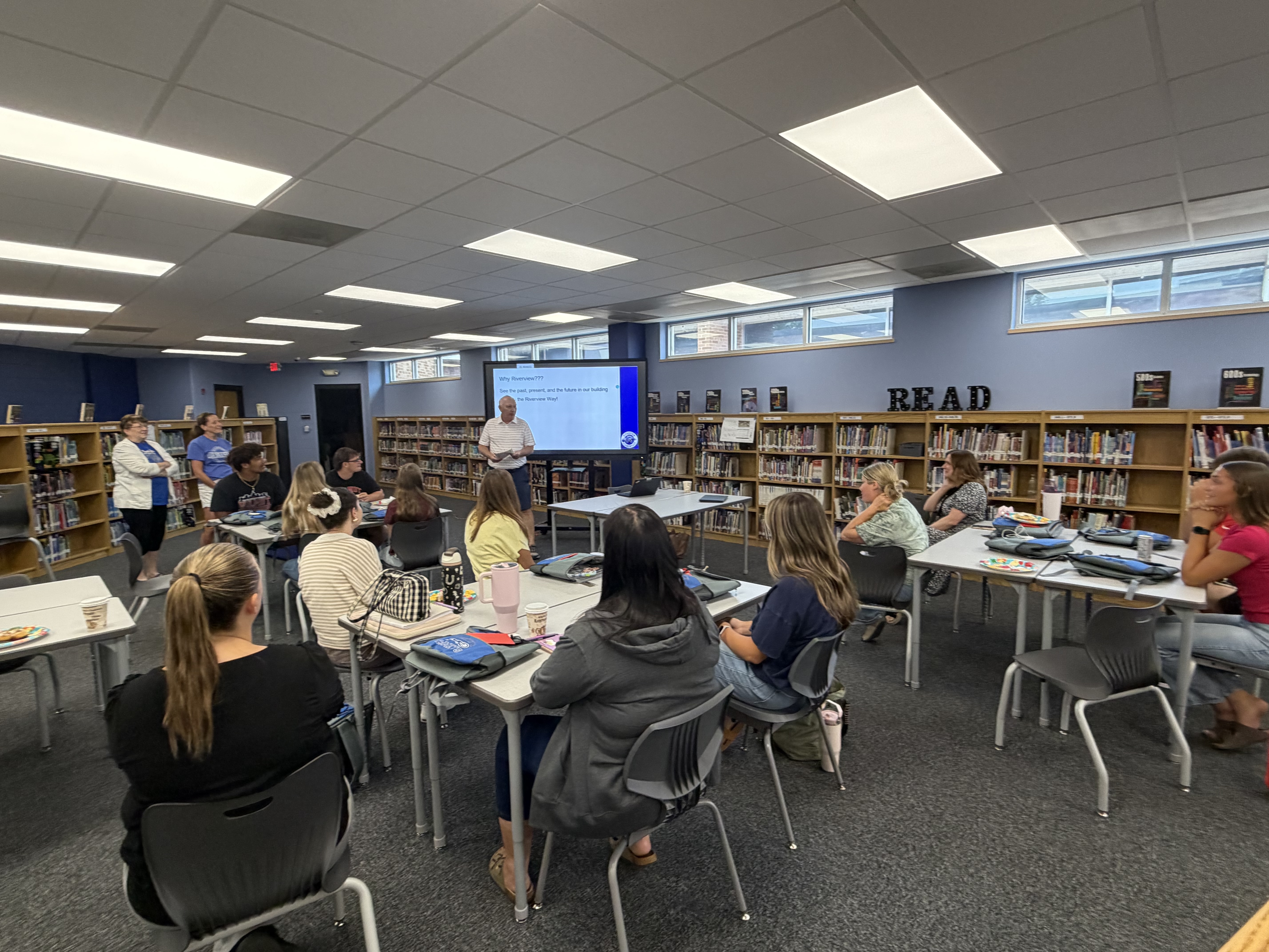 A presenter stands before a screen displaying 'Why Riverview??? See the past, present, and the future in our building the Riverview Way!' to an audience seated at tables.