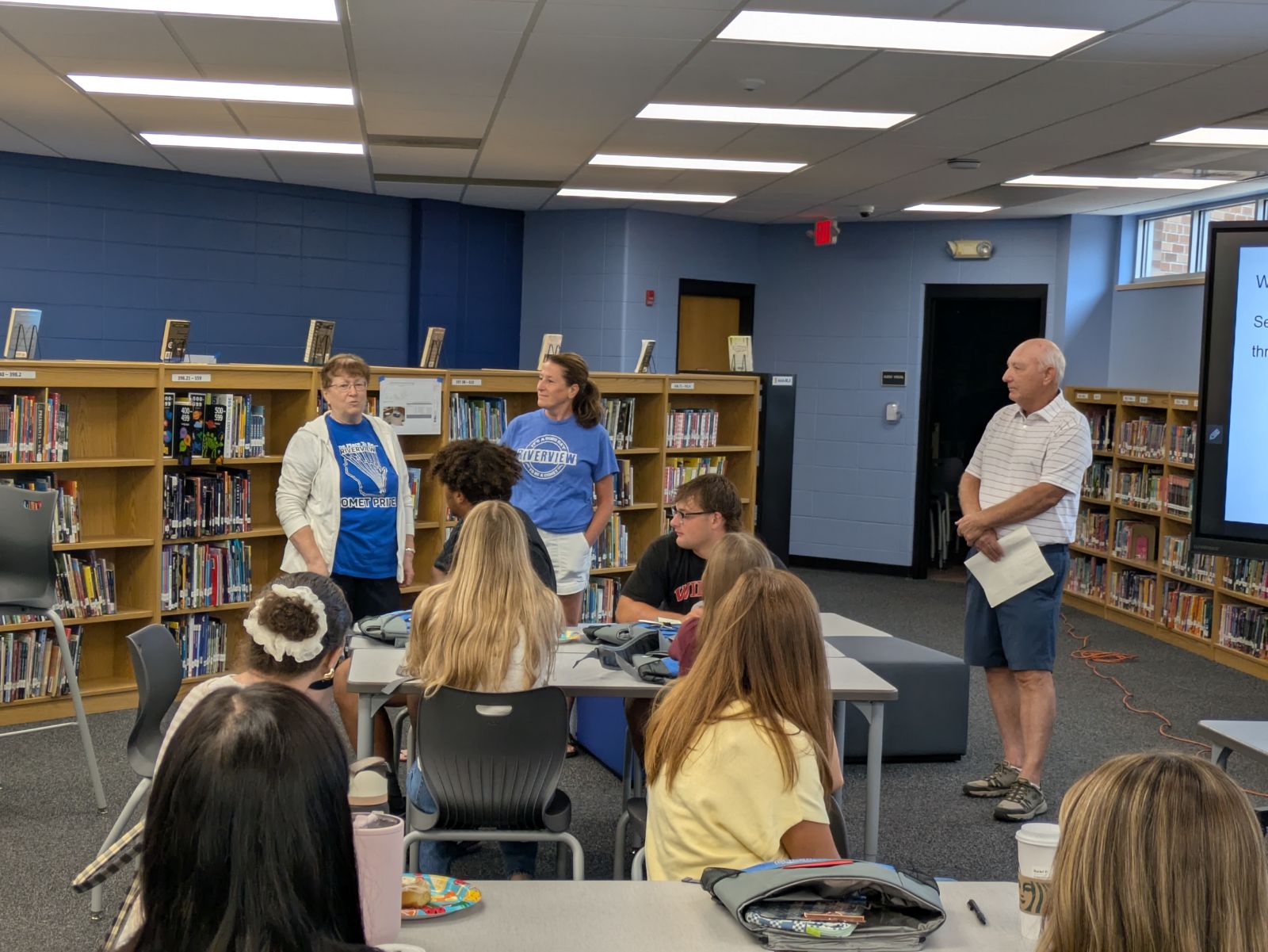 Two women in blue shirts and a man in a striped shirt stand in front of students in a library setting.