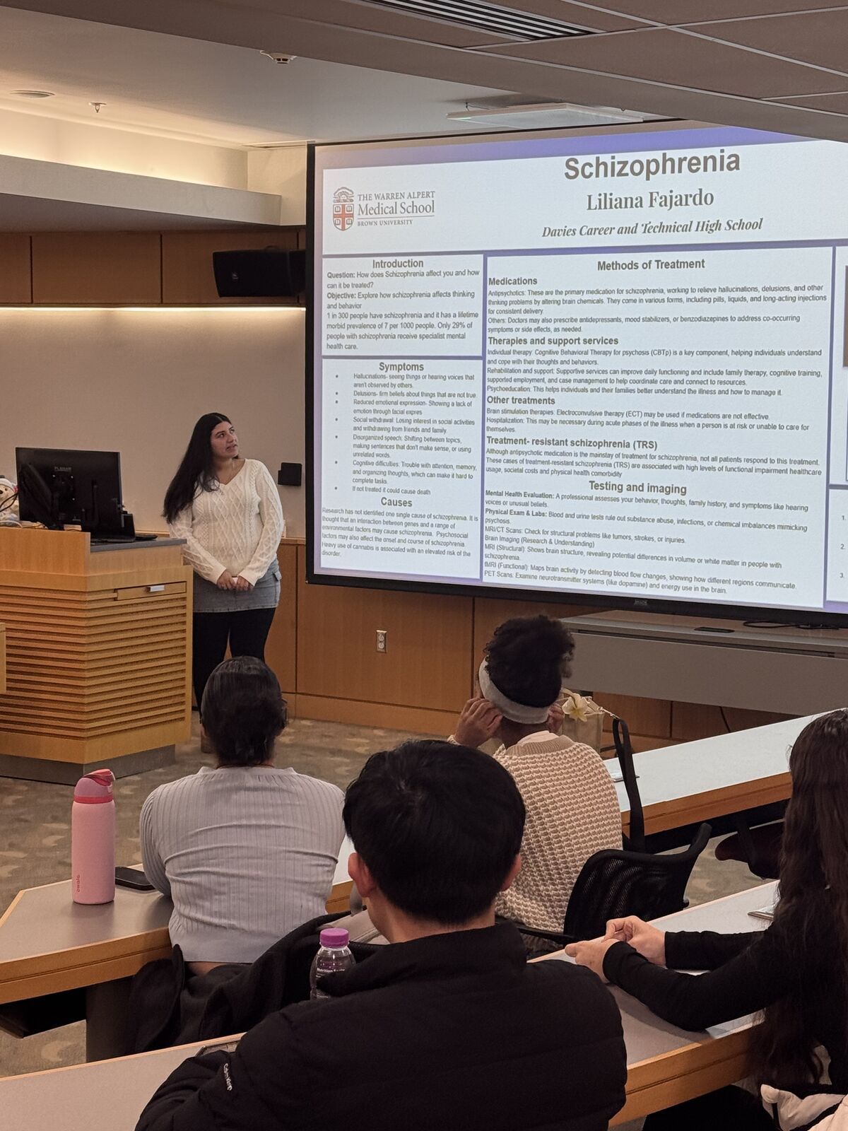  Lilliana stands near a large screen displaying information about Schizophrenia, with students seated in a classroom.