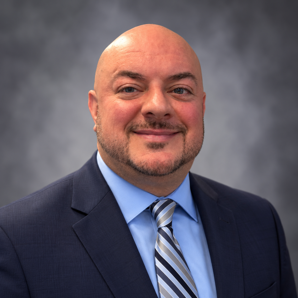 A professional headshot of a smiling man with a shaved head, wearing a dark suit, blue shirt, and striped tie.