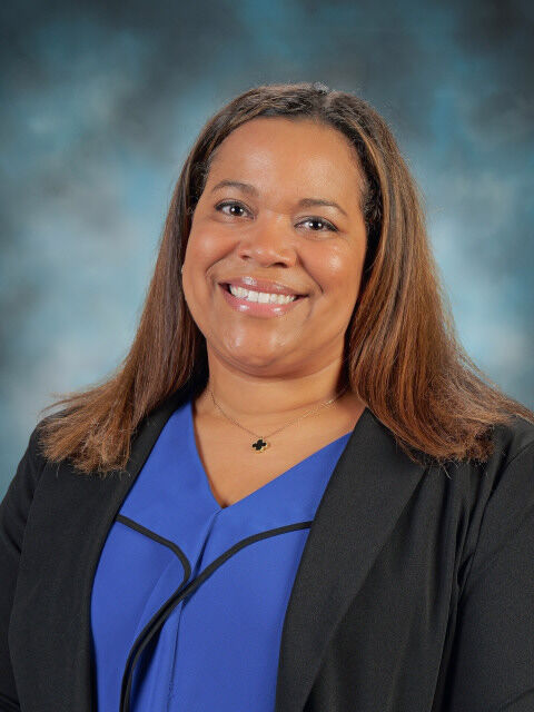 A professional portrait of a woman with long brown hair, wearing a blue top and a black blazer, smiling at the camera.