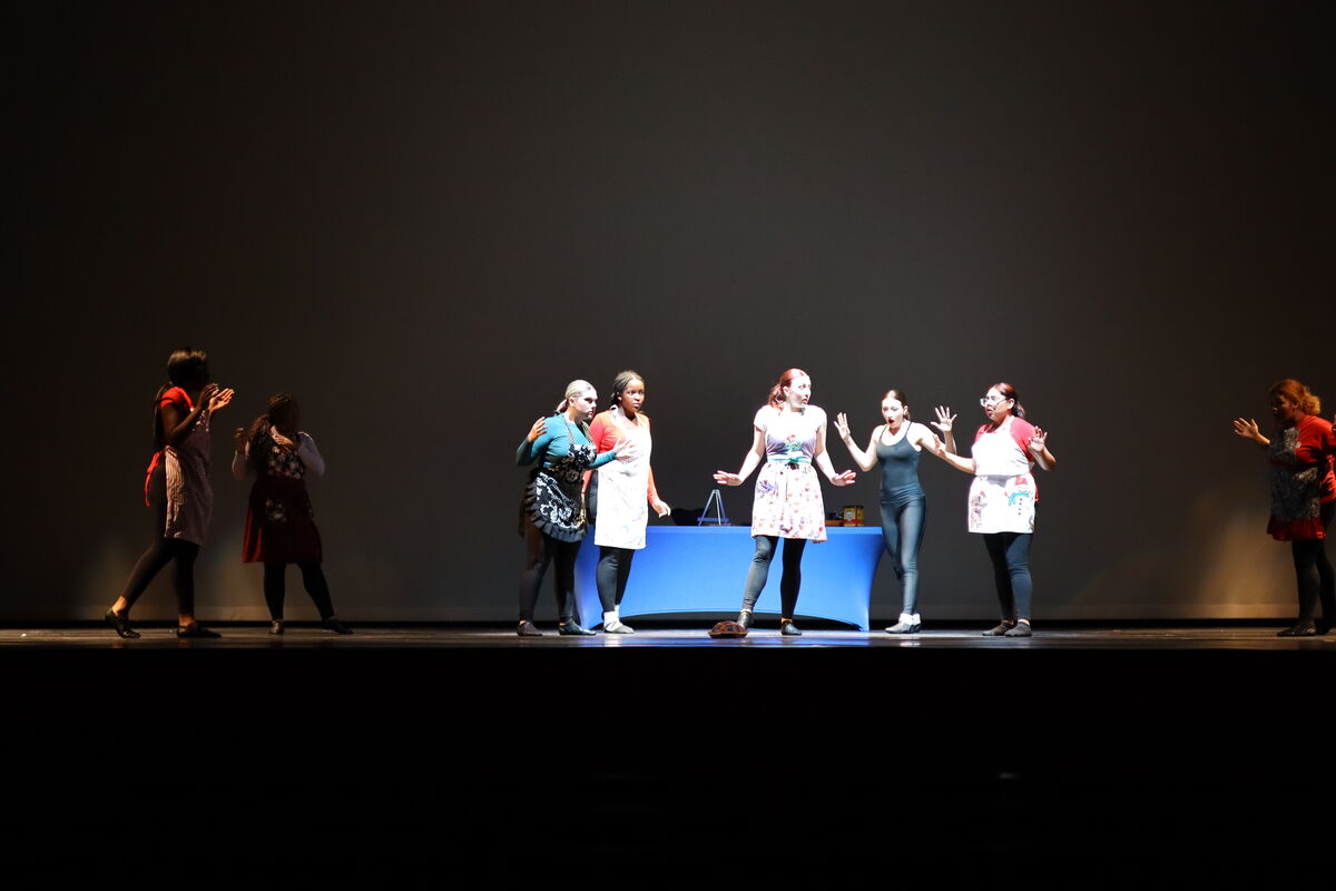 A group of women in various outfits, some wearing aprons, perform on a stage with a dark backdrop.