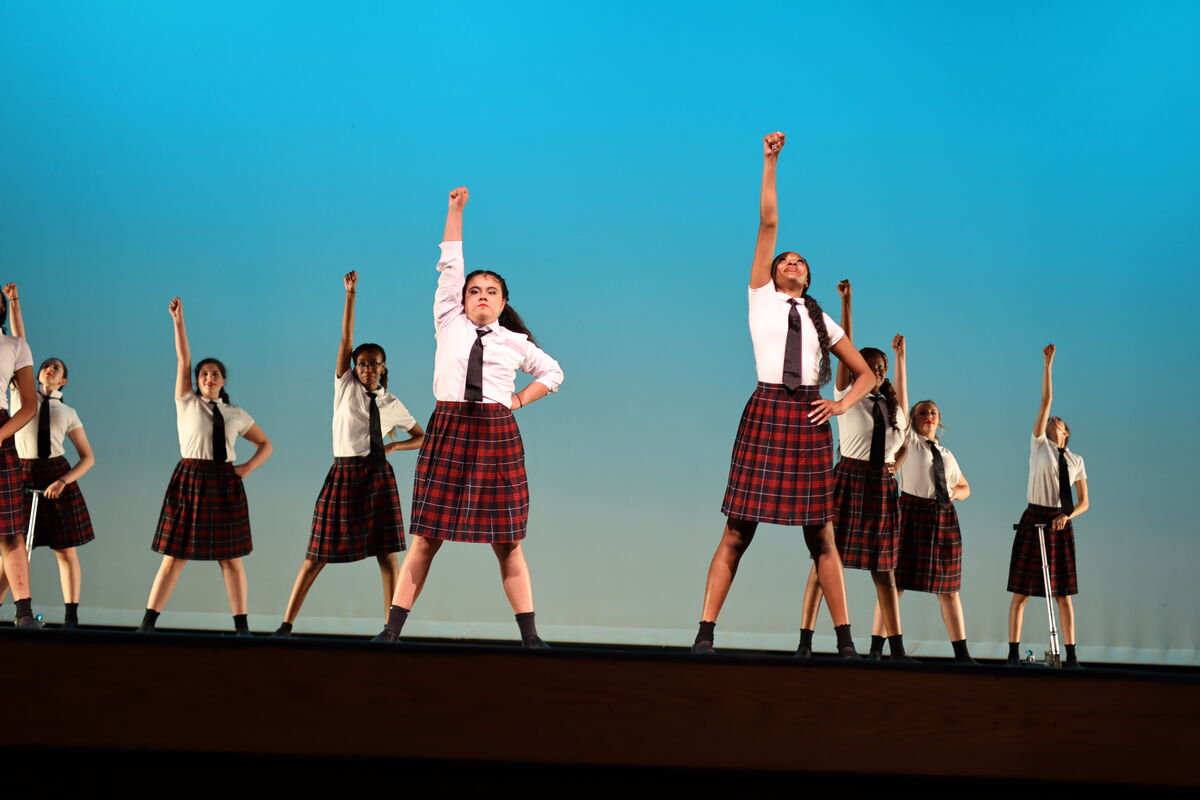 A group of women in various outfits, some wearing aprons, perform on a stage with a dark backdrop.