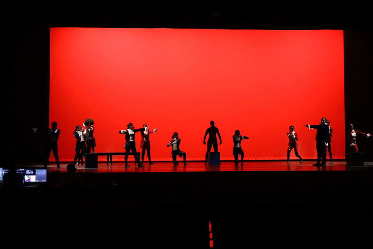 A group of women in various outfits, some wearing aprons, perform on a stage with a dark backdrop.