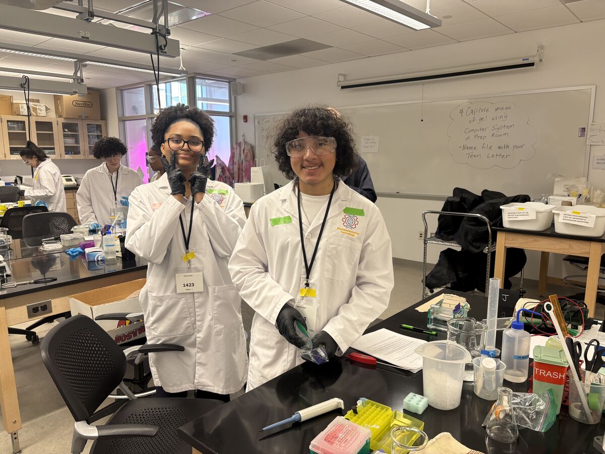 Two students in lab coats and safety goggles smile at the camera in a science lab, with lab equipment on the table.