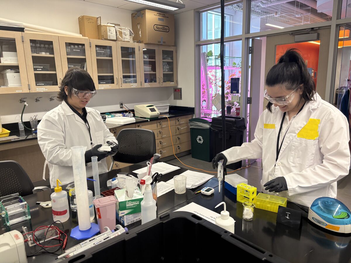 Two individuals in lab coats and safety glasses work at a lab bench with various scientific equipment, including beakers, test tubes, and a centrifuge.