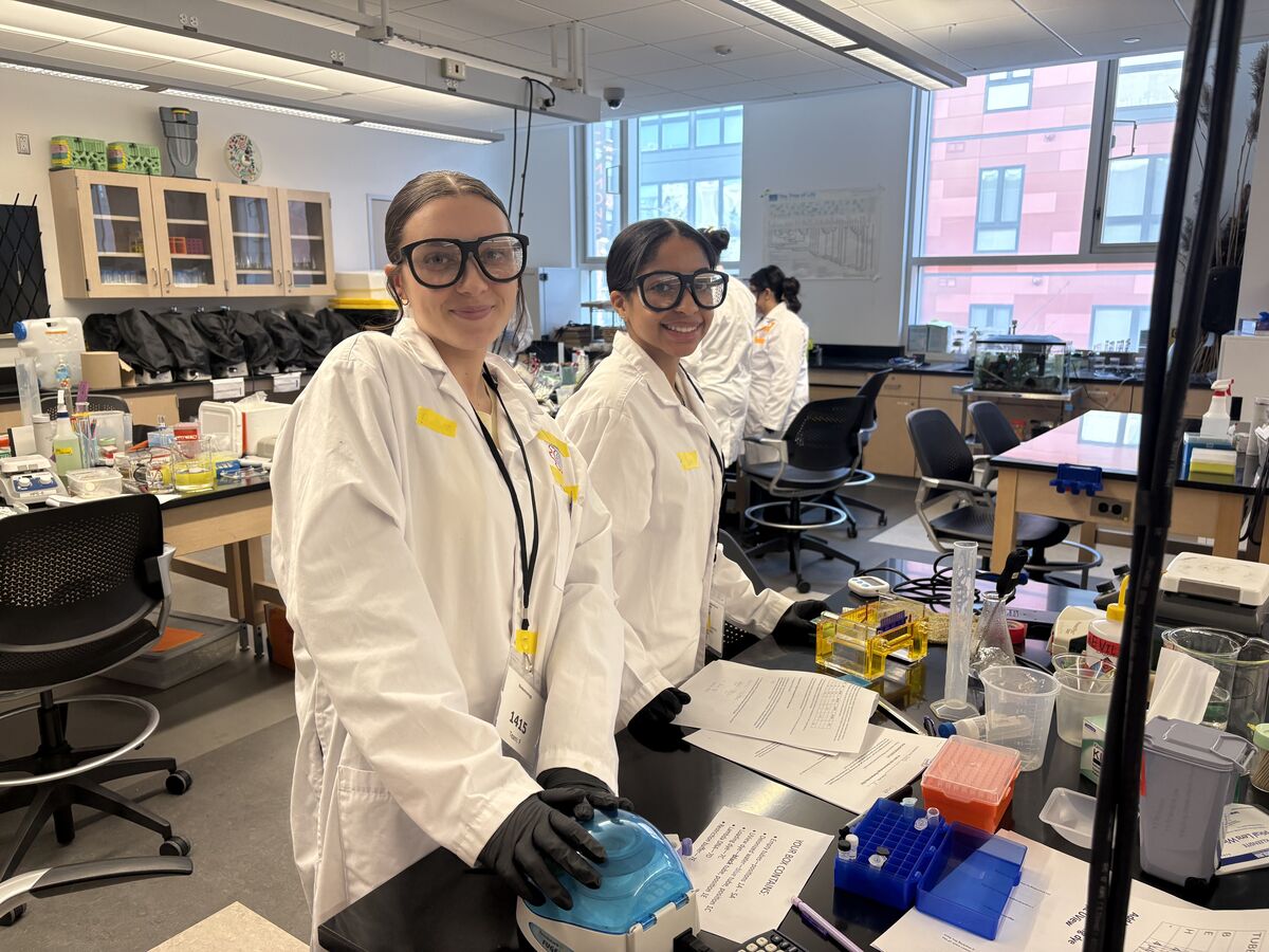 Two young women in lab coats and safety glasses smile while working in a laboratory setting.