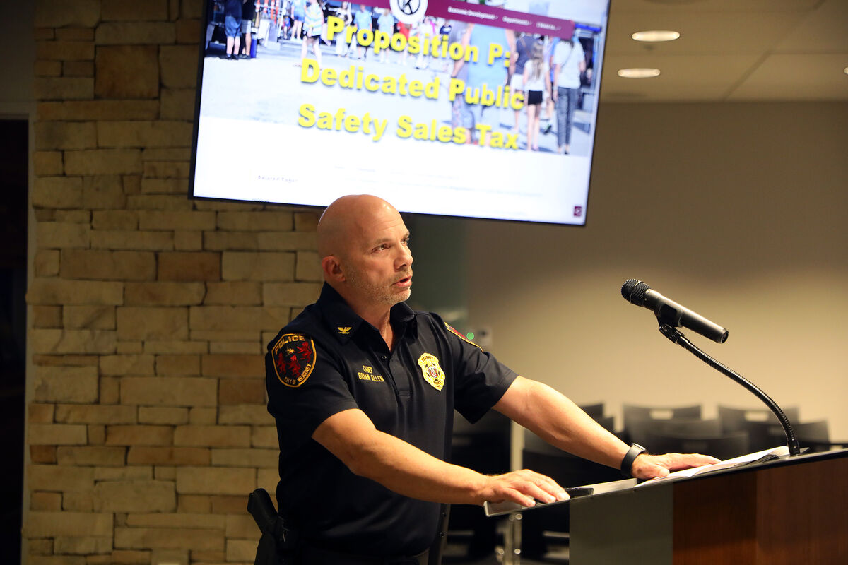 A police chief stands at a podium, speaking into a microphone with a screen displaying text behind him.