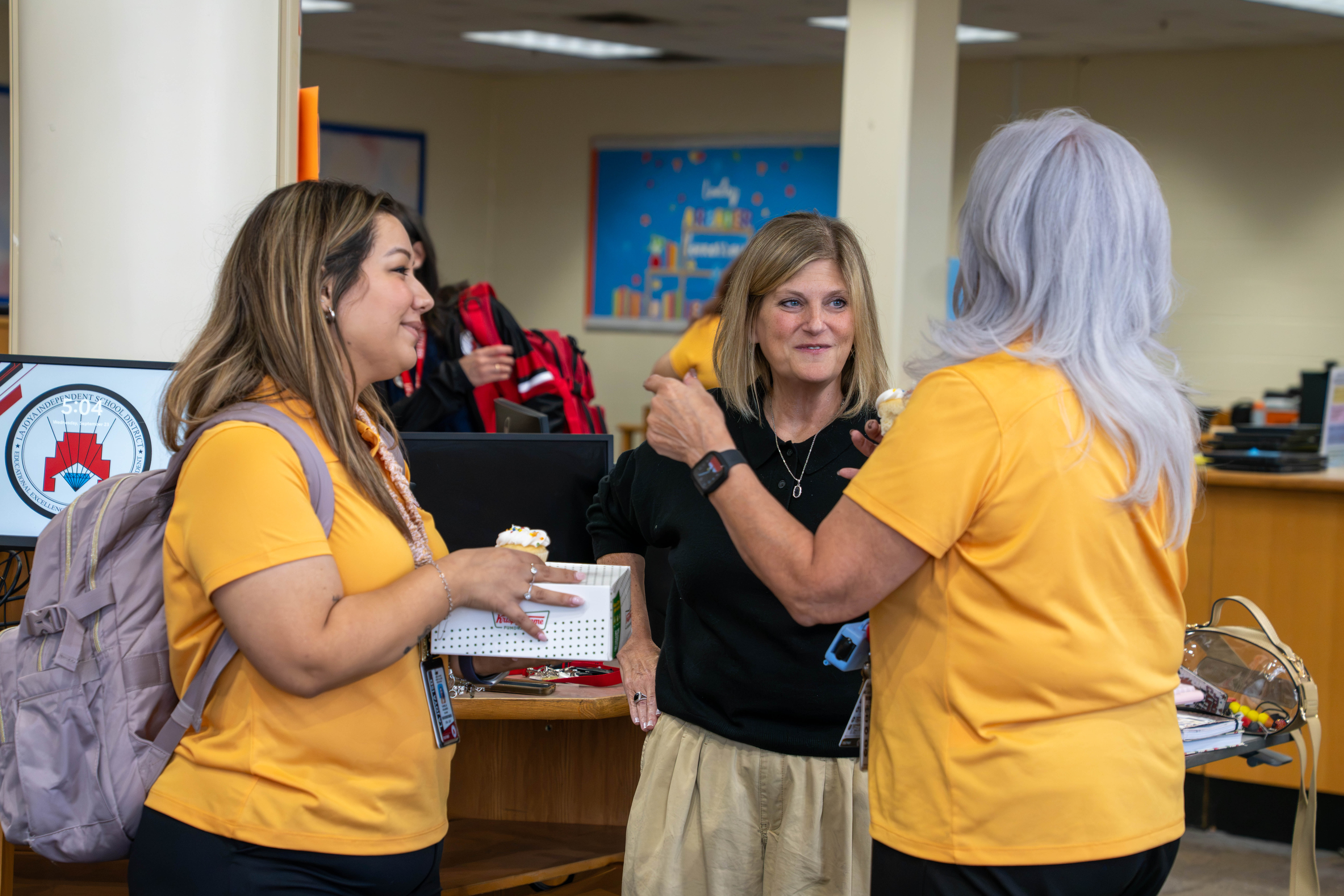 Three people converse and hold treats in an indoor setting, possibly an office or school.