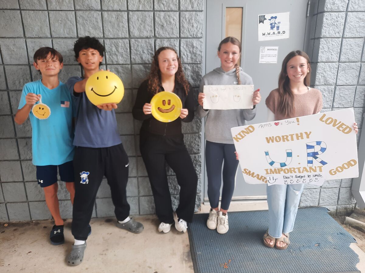 Five students pose with positive signs and smiley face decorations in front of a grey brick wall.