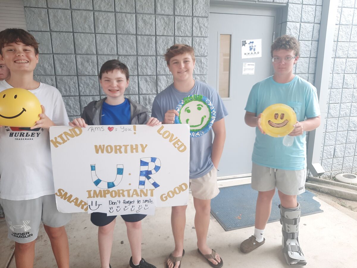 Four young people stand outside a building, holding positive signs and smiley face crafts.