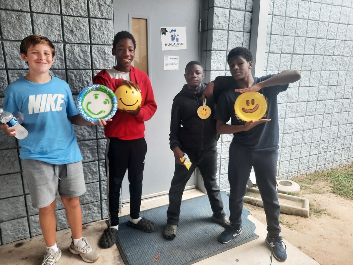 Four smiling youths pose outdoors holding smiley face paper plates and decorations, standing near a grey block wall.