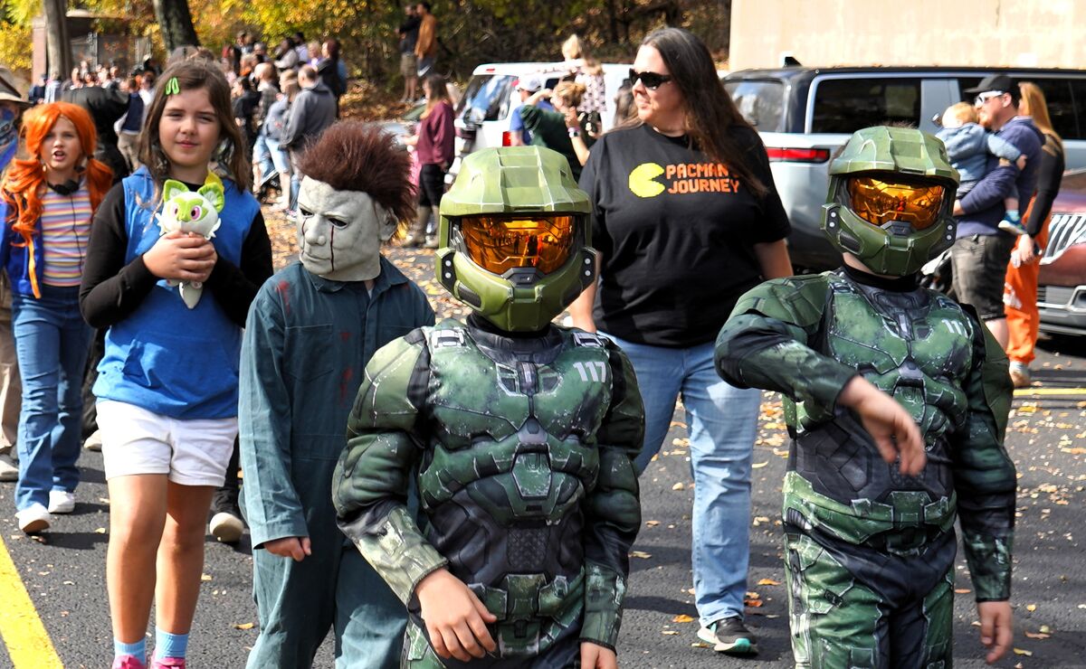 A group of children and adults in costumes, including two in Master Chief armor, walk down a sunny street.