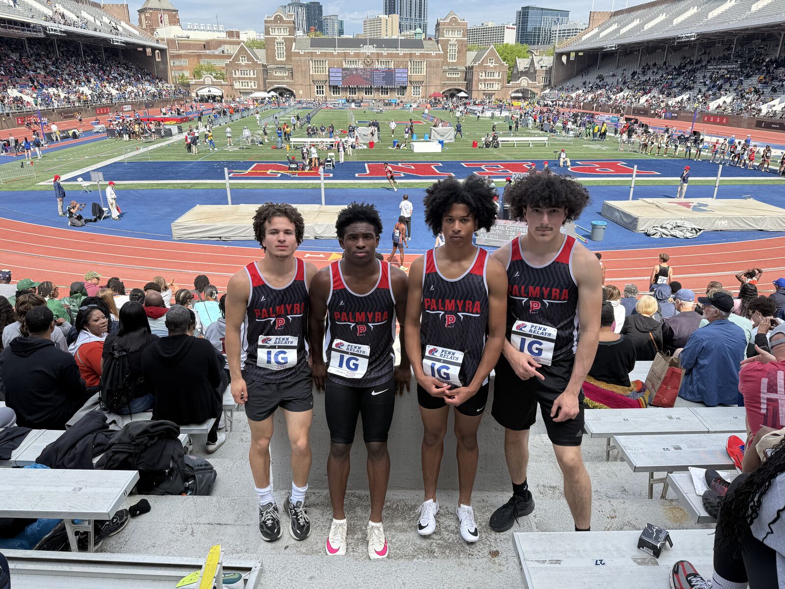 Four young male athletes in matching Palmyra track uniforms stand together at Penn Relays stadium.