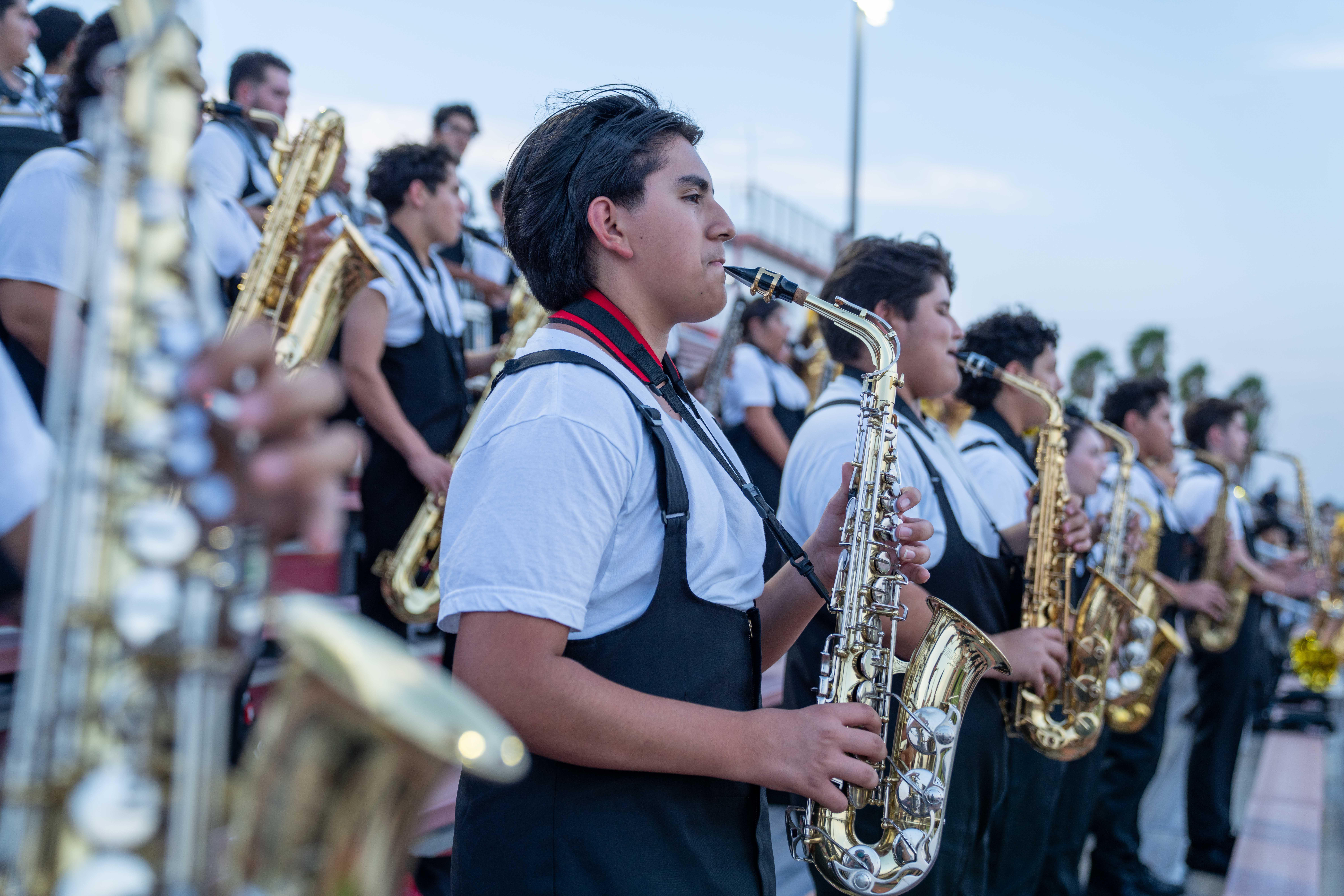 A marching band saxophonist plays an instrument outdoors, focused on the music.