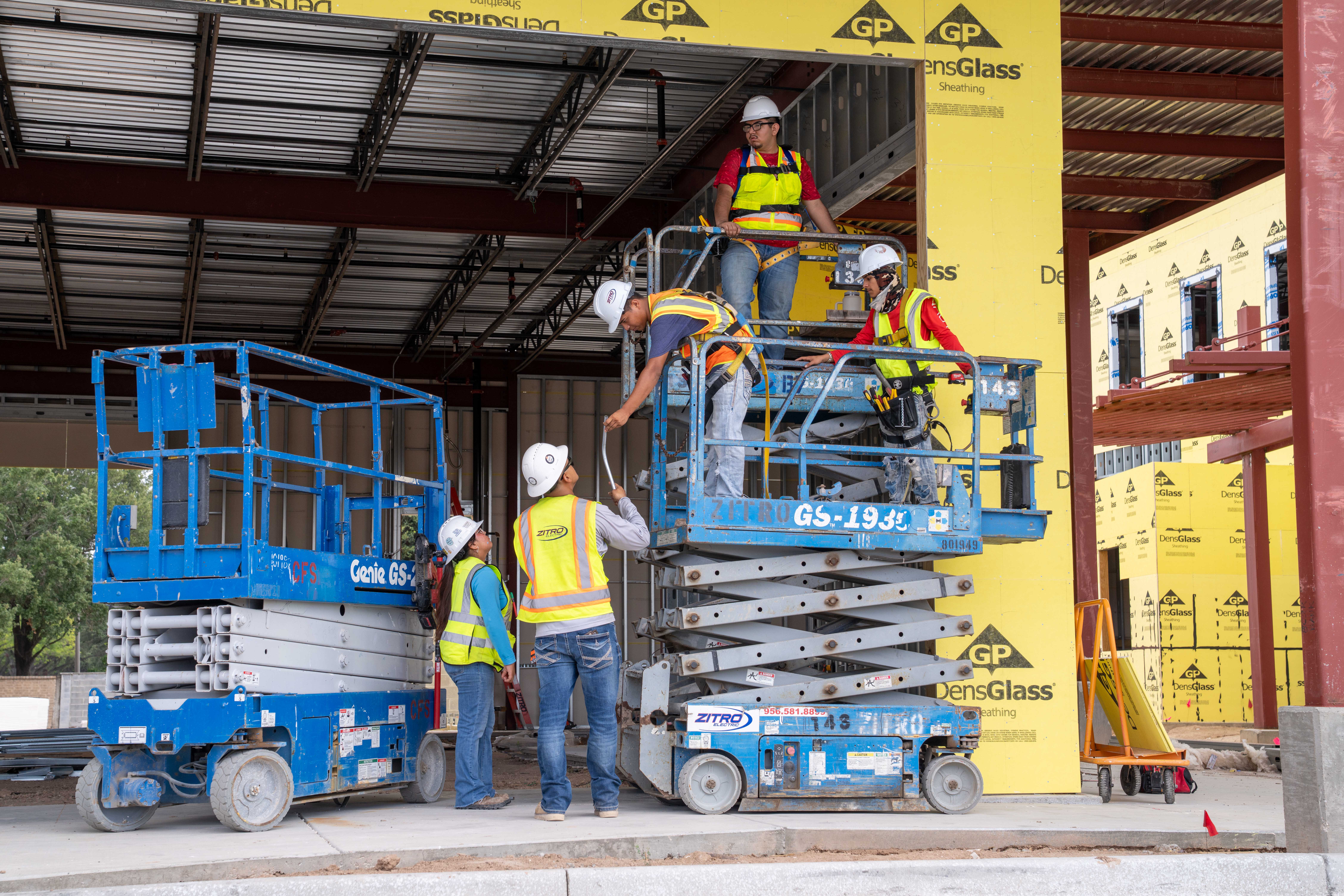 Construction workers in hard hats and safety vests operate a scissor lift on a building site.