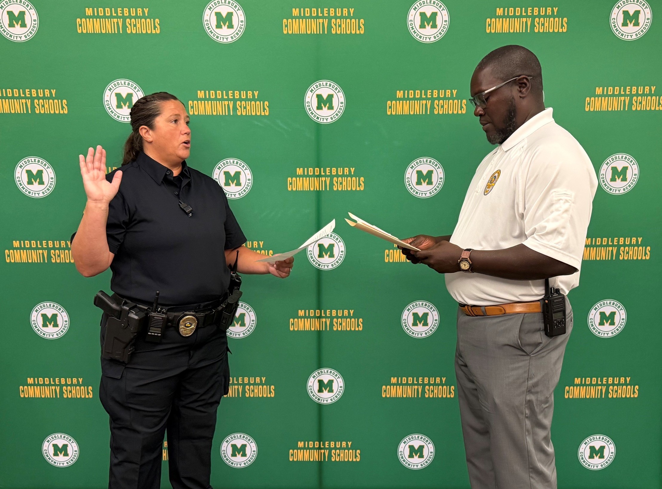 A woman in a police uniform raises her hand while holding papers, facing a man in a polo shirt who is also holding papers.