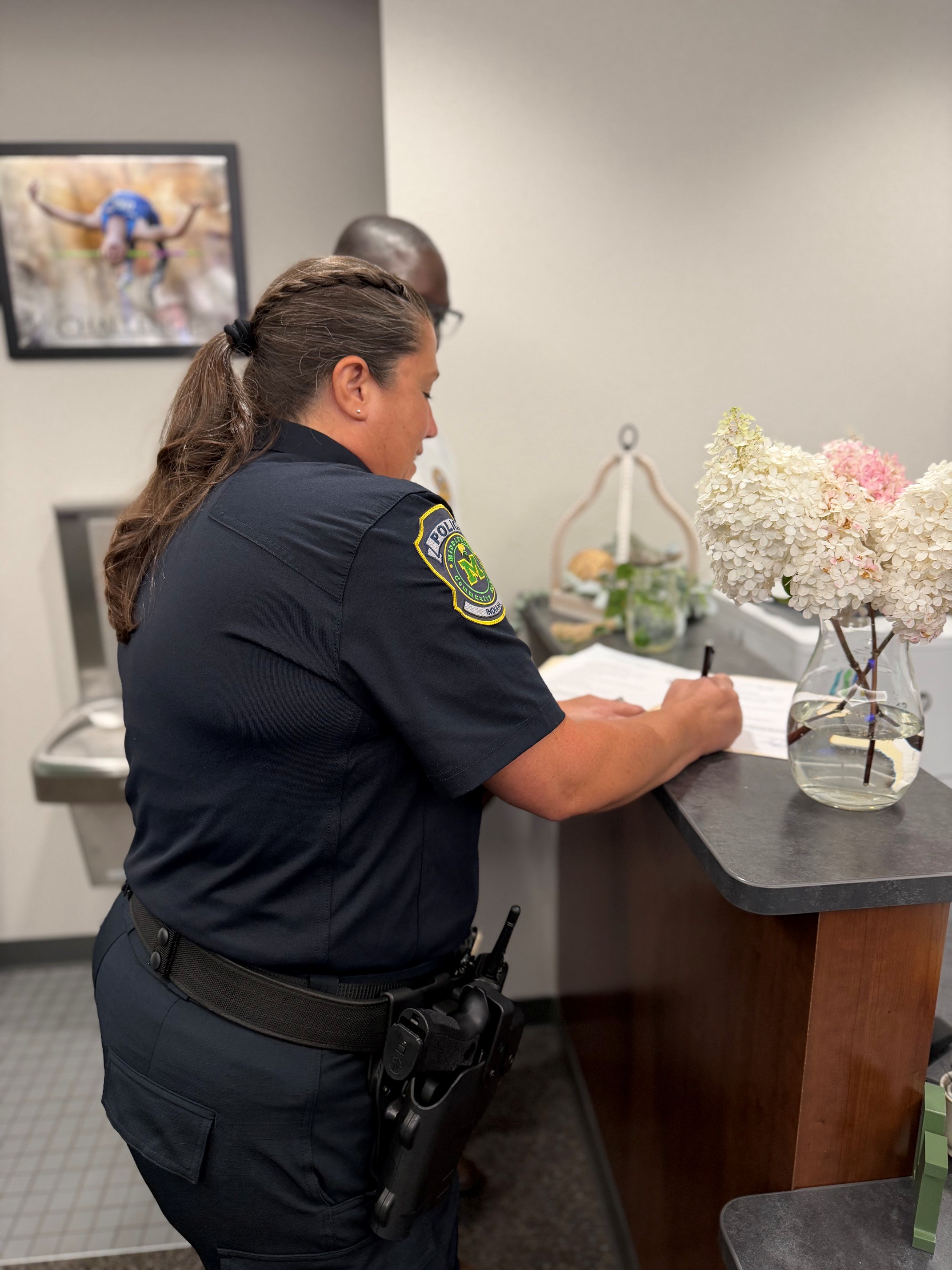 A female officer in uniform writes on a document at a counter, with a vase of flowers nearby.