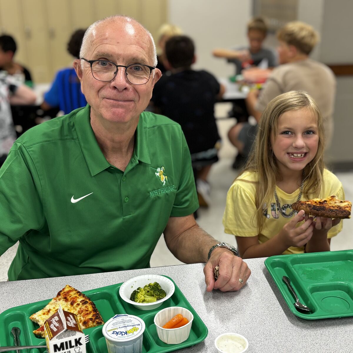 A man and a young girl smile at the camera while sitting at a school cafeteria table with lunch trays.