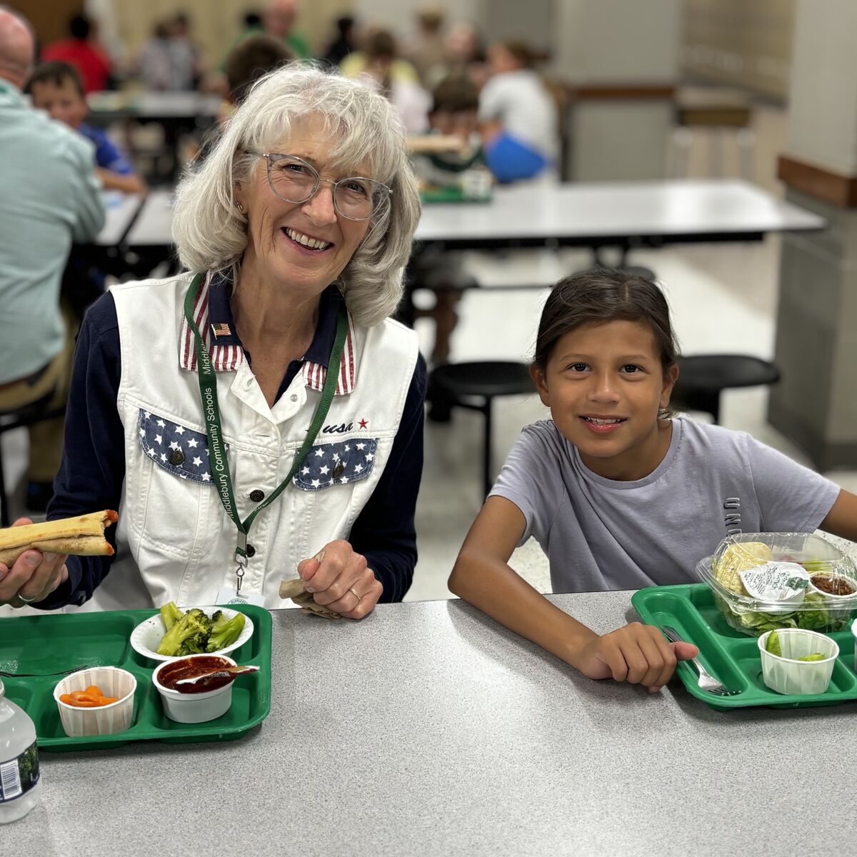 A woman and a young girl smile while sitting at a table with lunch trays in a cafeteria.