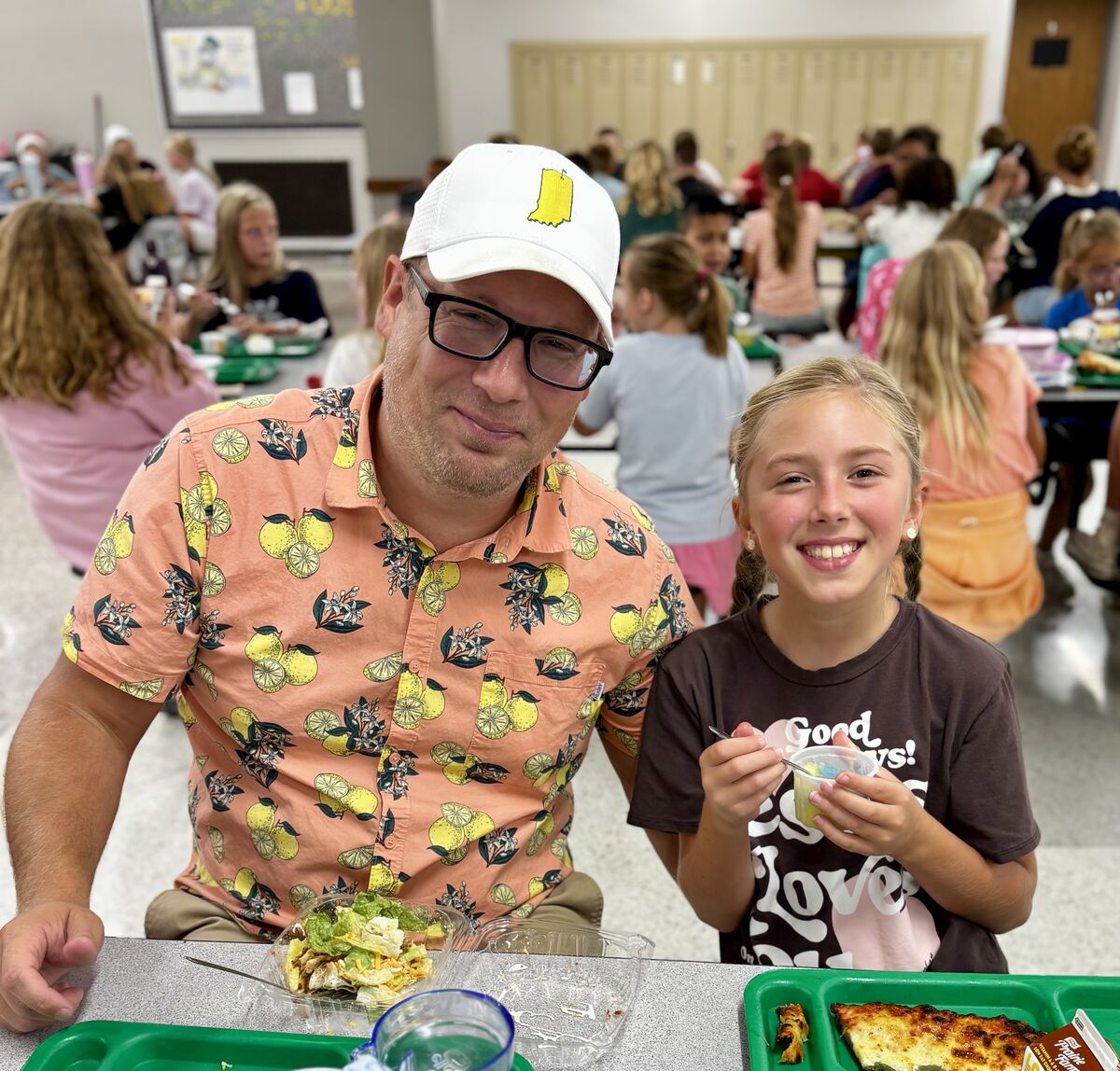 A man and a young girl smile at the camera while sitting at a school cafeteria table.
