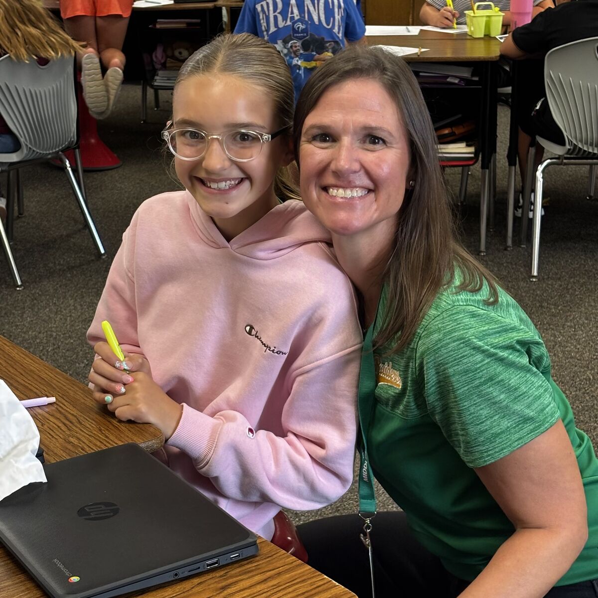 A young student with glasses and a pink hoodie smiles next to a woman in a green shirt.