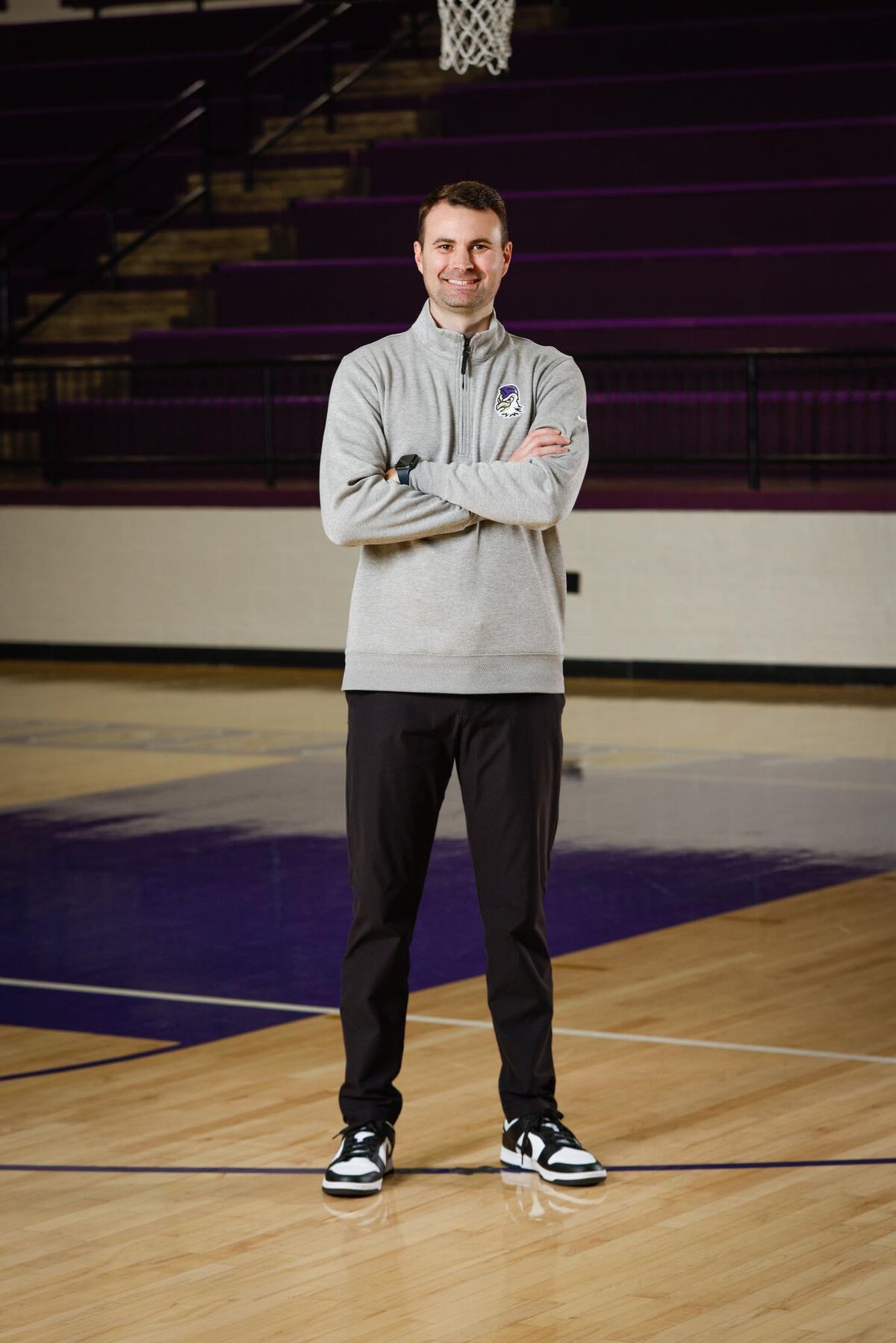 A man in a grey quarter-zip sweatshirt and black pants stands on a basketball court with his arms crossed.
