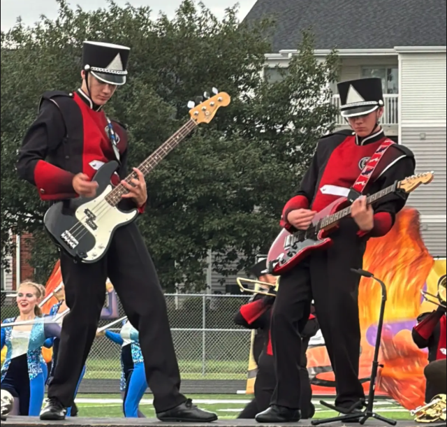 Two marching band members in red and black uniforms play electric guitars outdoors.