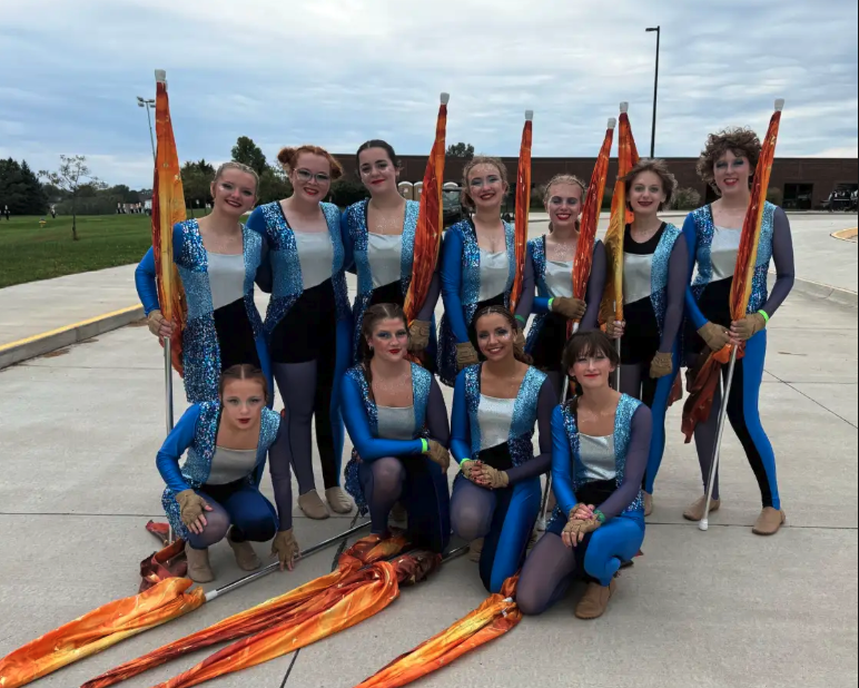 A group of ten color guard members pose together outdoors, holding orange and yellow flags and wearing matching blue and silver costumes.