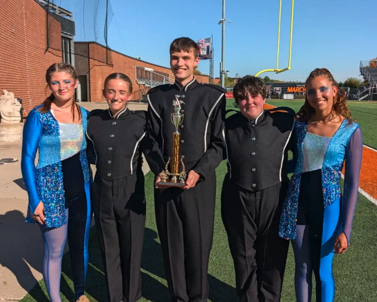 Five high school students in marching band uniforms and performance attire proudly pose with a trophy outdoors.