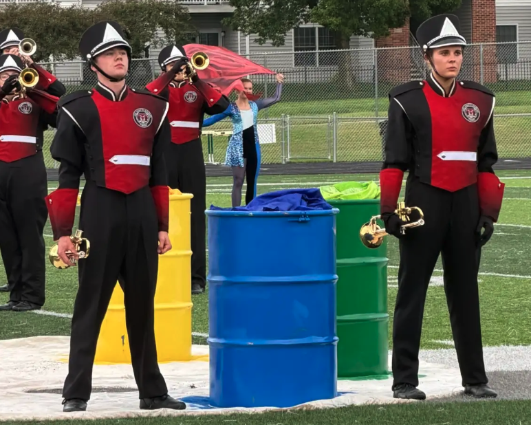 Marching band members in red, black, and white uniforms stand on a field with colorful barrels.