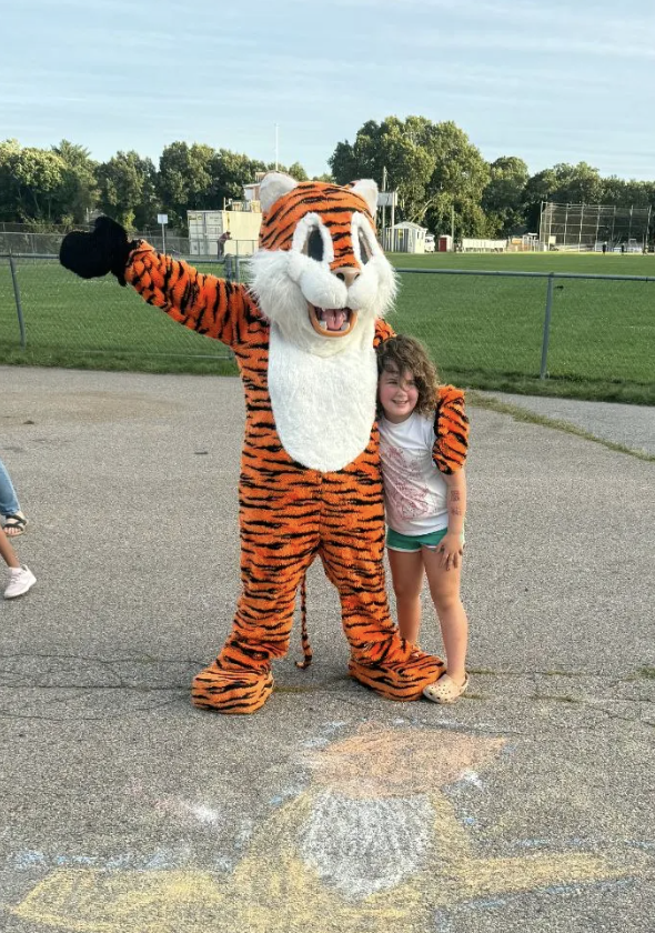A child smilingly hugs a friendly tiger mascot outdoors near a grassy field.