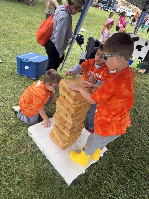 Three children in matching orange shirts play with a large wooden block tower on a grass lawn.