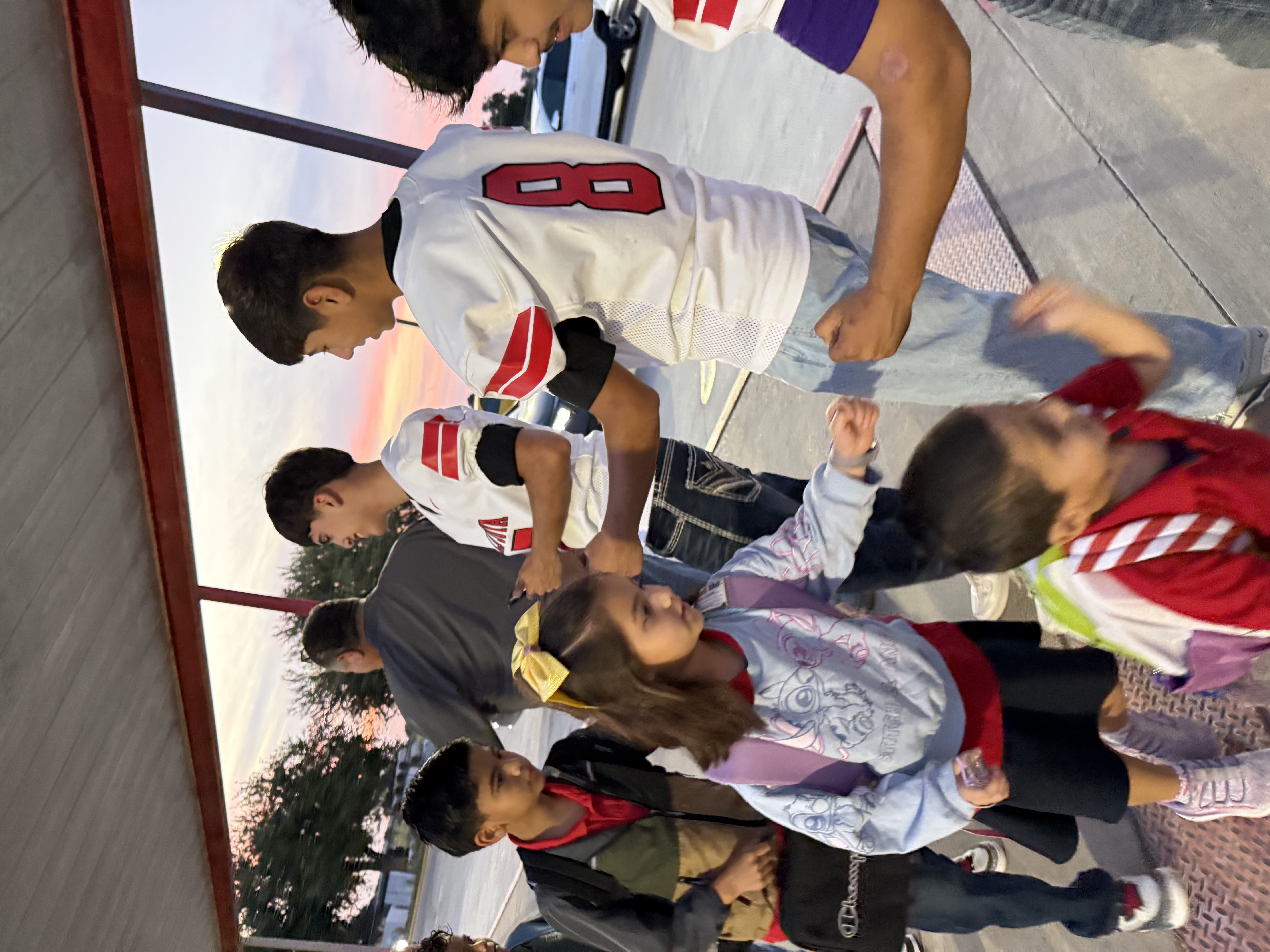 A group of people, including young adults in football jerseys and children, gather outside during sunset.