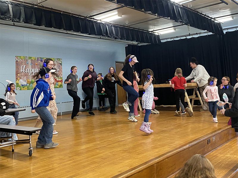 A group of children and adults wearing headphones are dancing on a wooden stage.