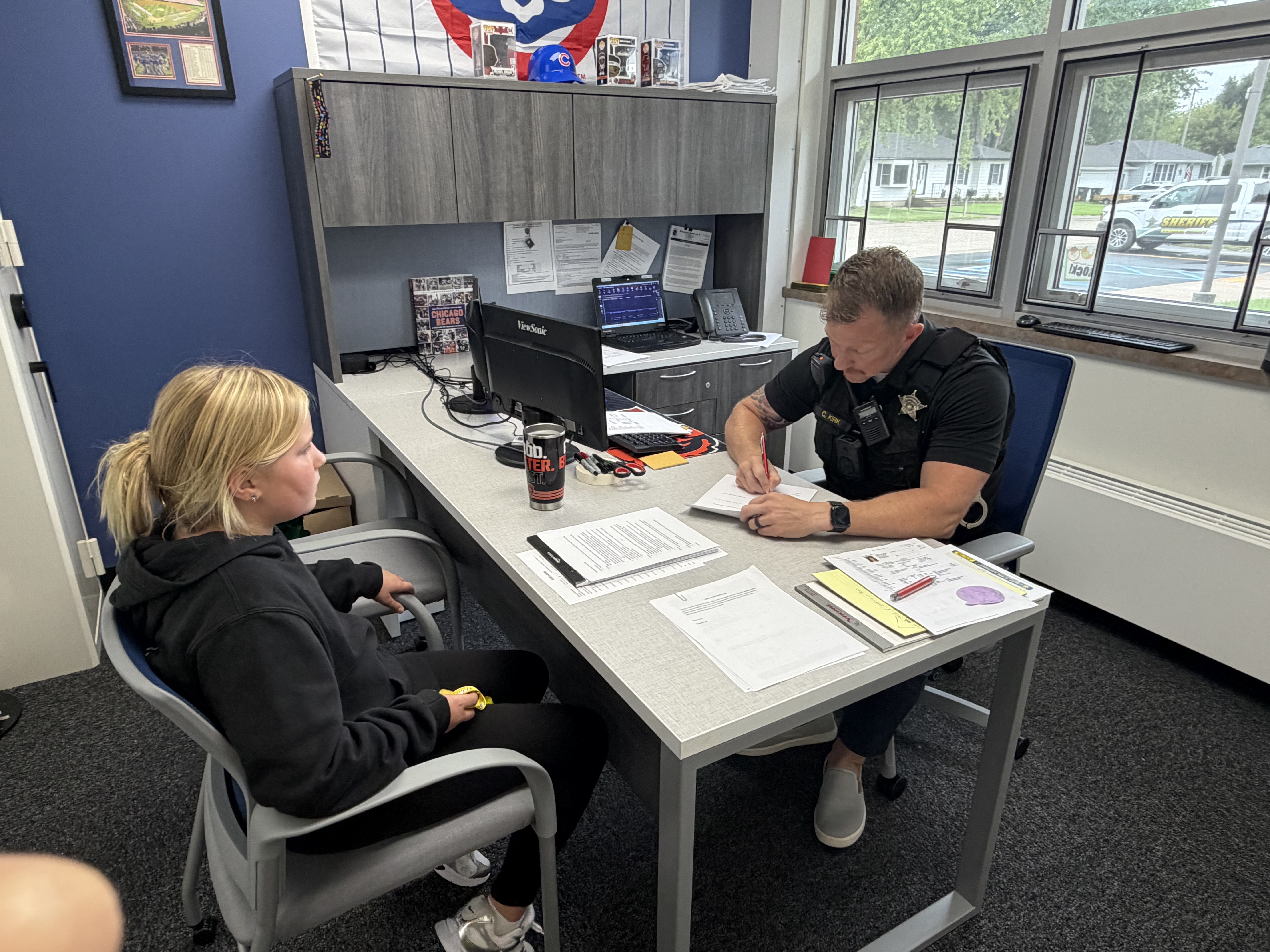 A young girl sits across from a police officer at a desk in an office.