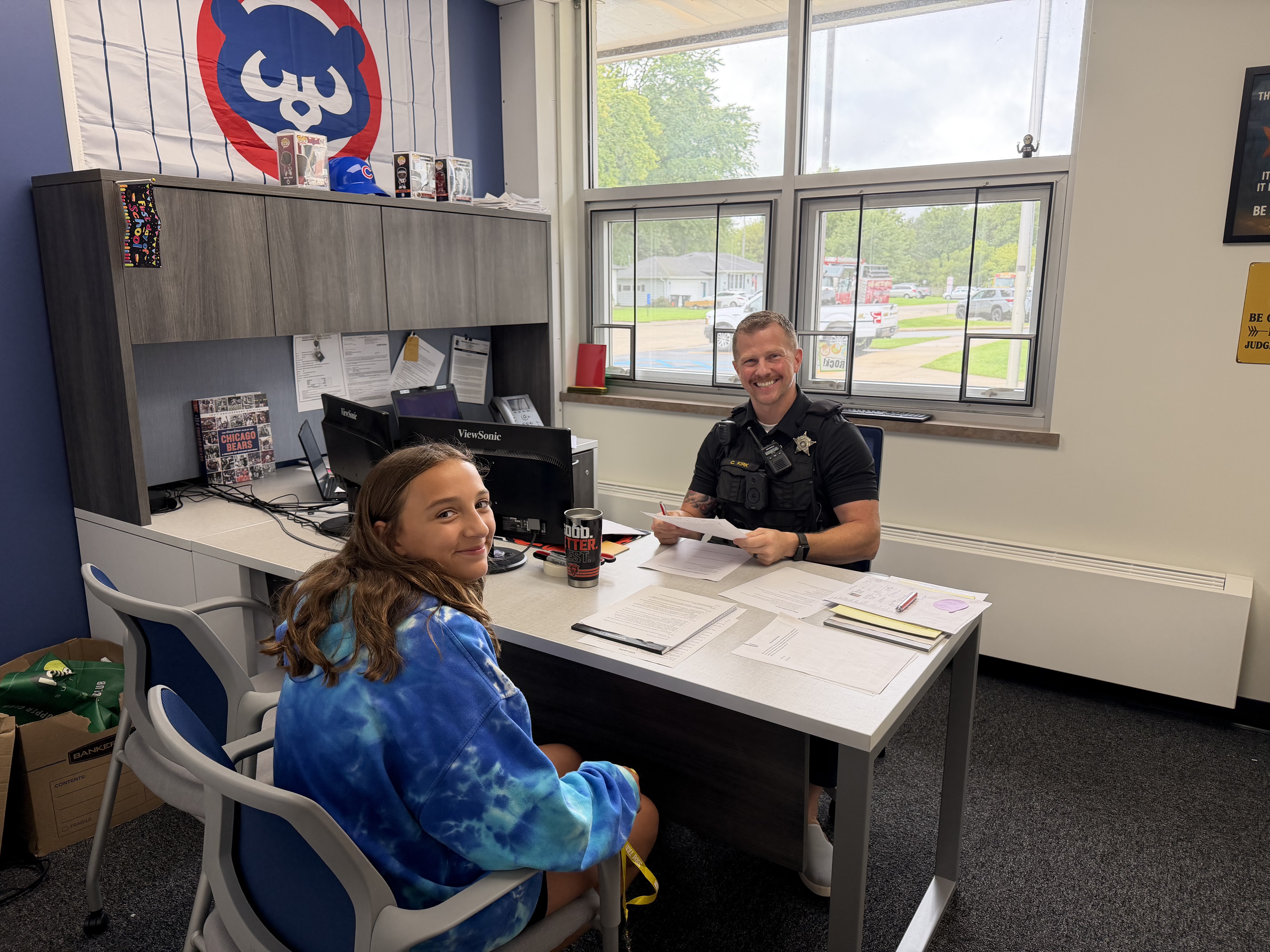 A smiling police officer sits at a desk across from a young person in an office setting with a Chicago Cubs flag on the wall.