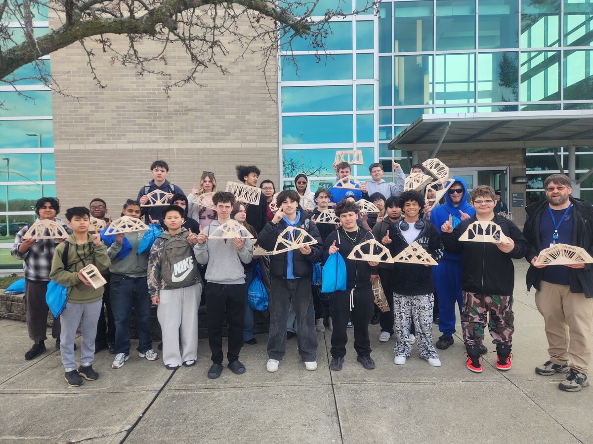 A diverse group of students proudly displays their handmade wooden bridge models outside a modern building.