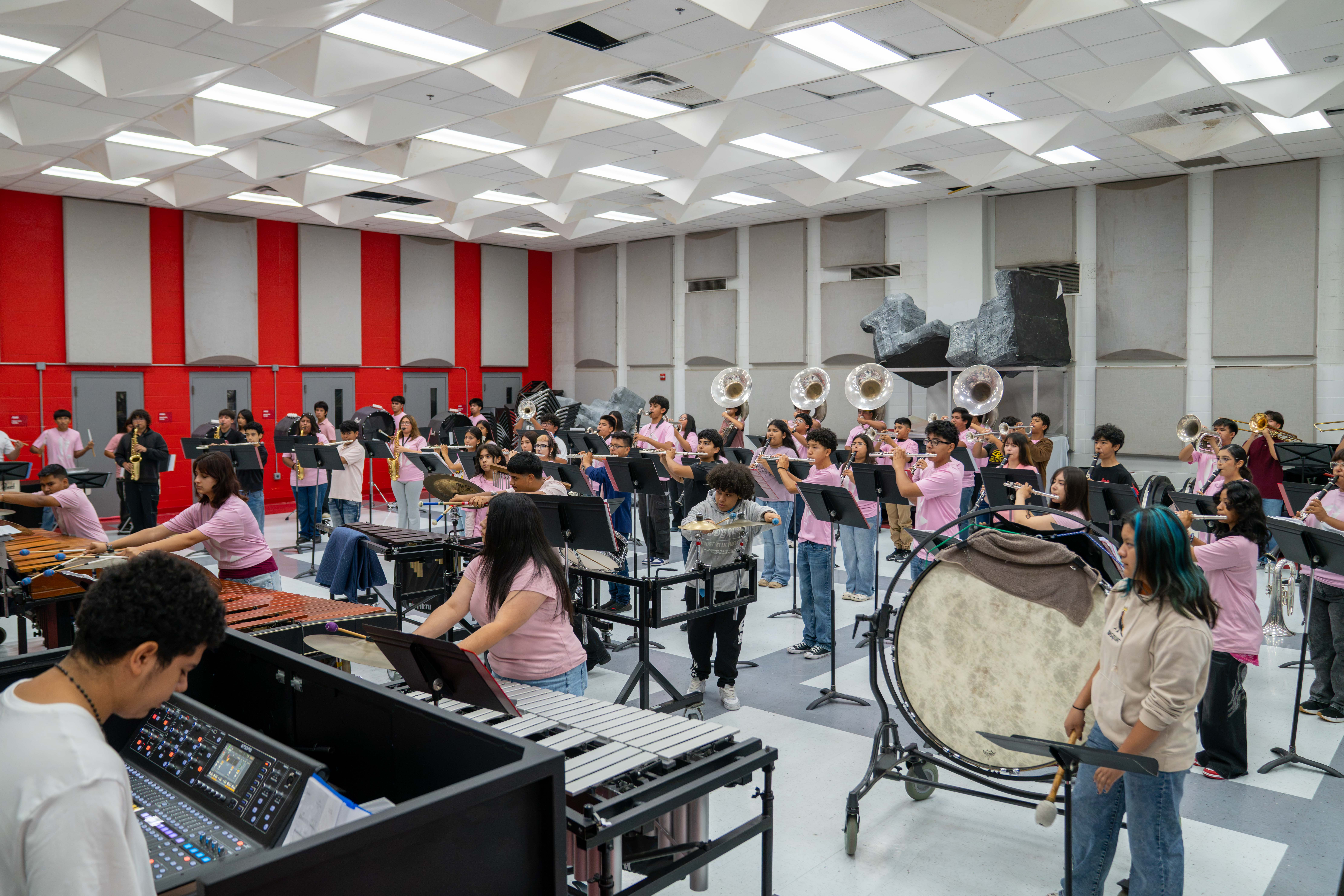 A large school band practices inside a brightly lit rehearsal room with red and white acoustic panels.