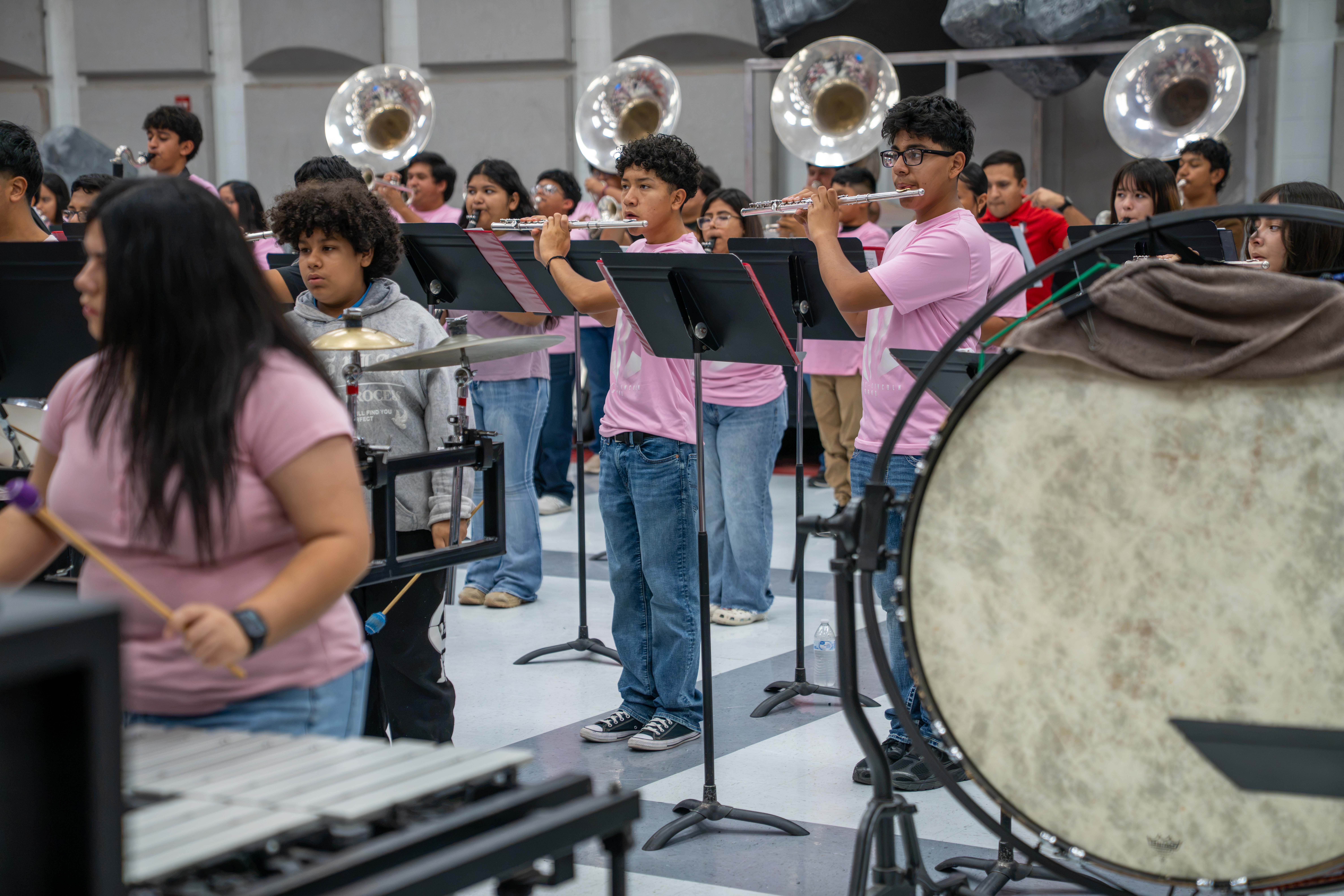 A high school marching band practices indoors, with students playing flutes, percussion, and brass instruments.
